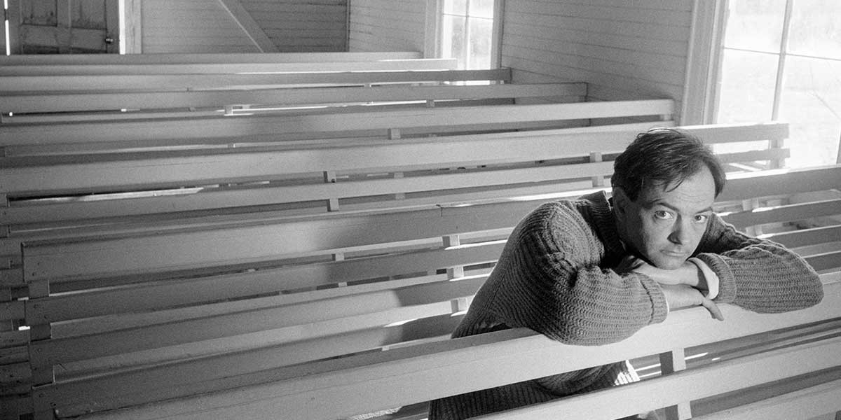 Man in a thick sweater resting his arms and head on a bench in an empty sunlit room with wooden benches.