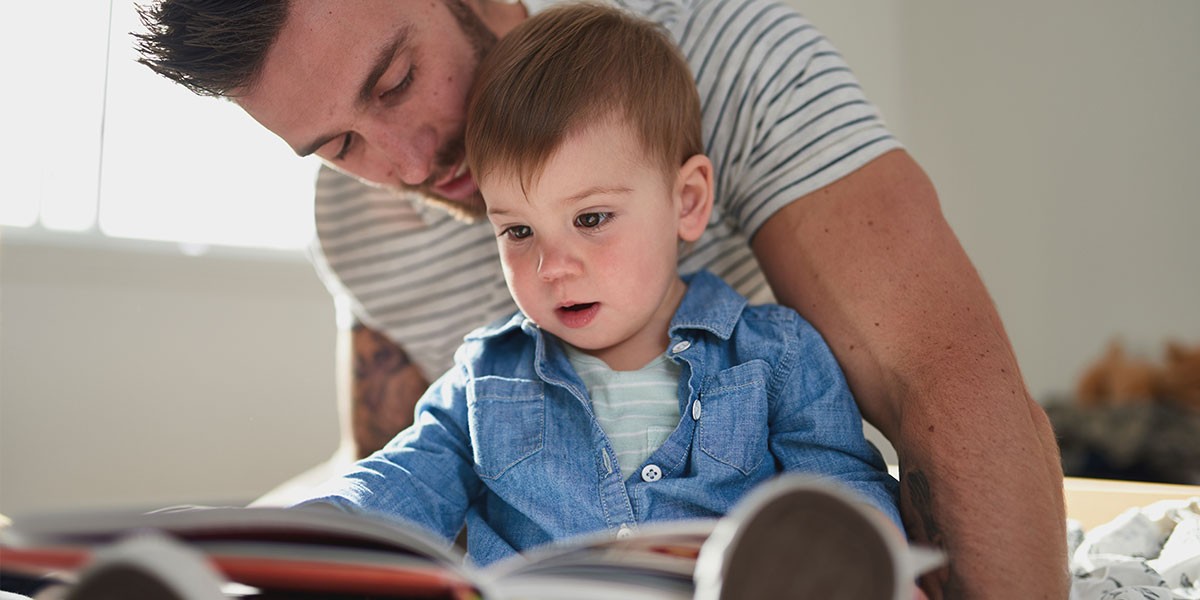 Man in striped shirt reading a book with a toddler wearing a blue shirt on his lap.