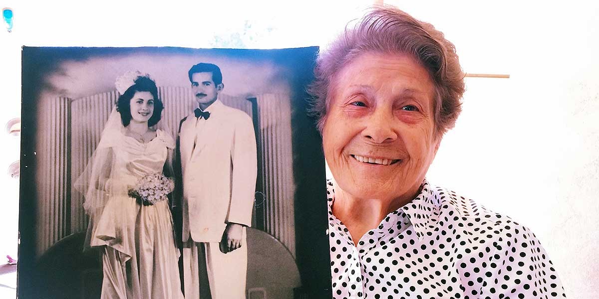 Elderly woman smiling and holding a vintage black and white wedding photo of a bride and groom.