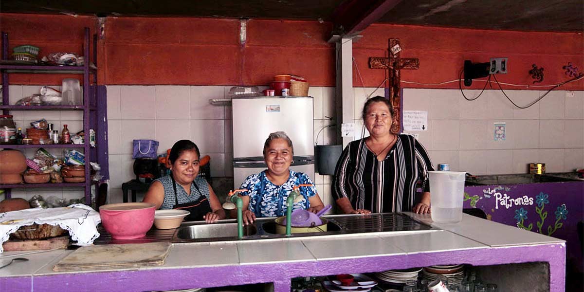 Three women standing behind a kitchen counter with shelves, bowls, and a refrigerator behind them.