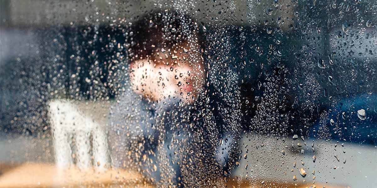 Person sitting indoors behind a rain-covered window, visible through water droplets.