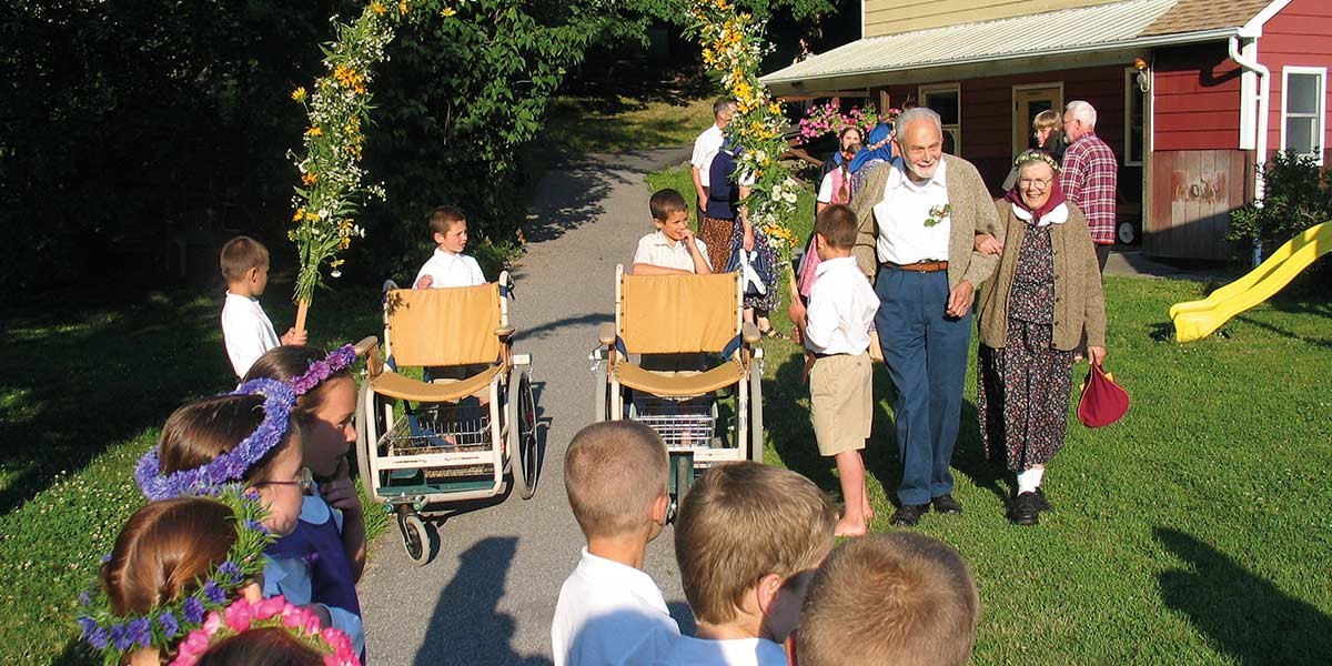 Children in white shirts with flower crowns and decorated wheelchairs walking outdoors, with elderly couple holding hands.