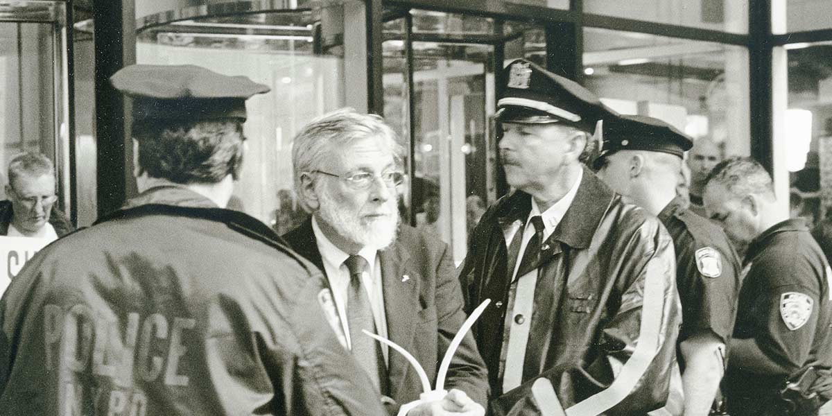 Black and white photo of a man with glasses and beard speaking to uniformed police officers near glass doors.