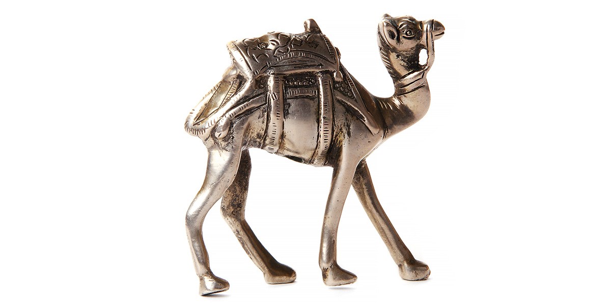 Small metallic figurine of a camel with detailed saddle and harness walking on four legs.