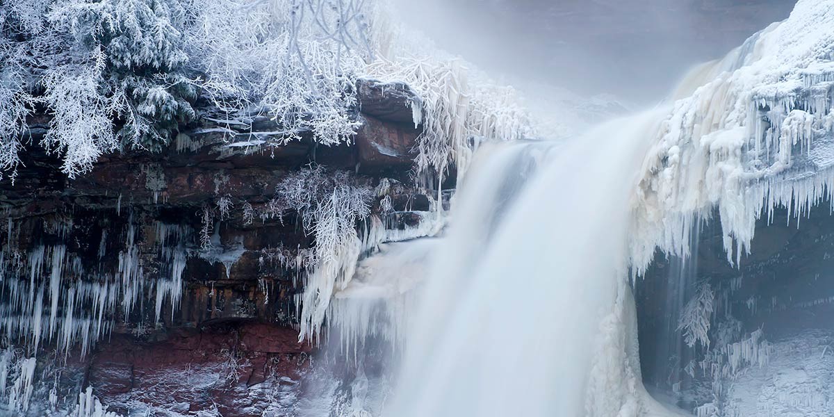 Waterfall flowing beside a cliff with trees and rocks covered in ice and snow in winter.