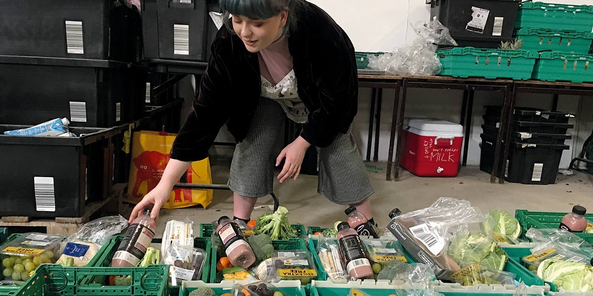 Person organizing crates with broccoli, grapes, juice bottles, and packaged vegetables in a storage room.