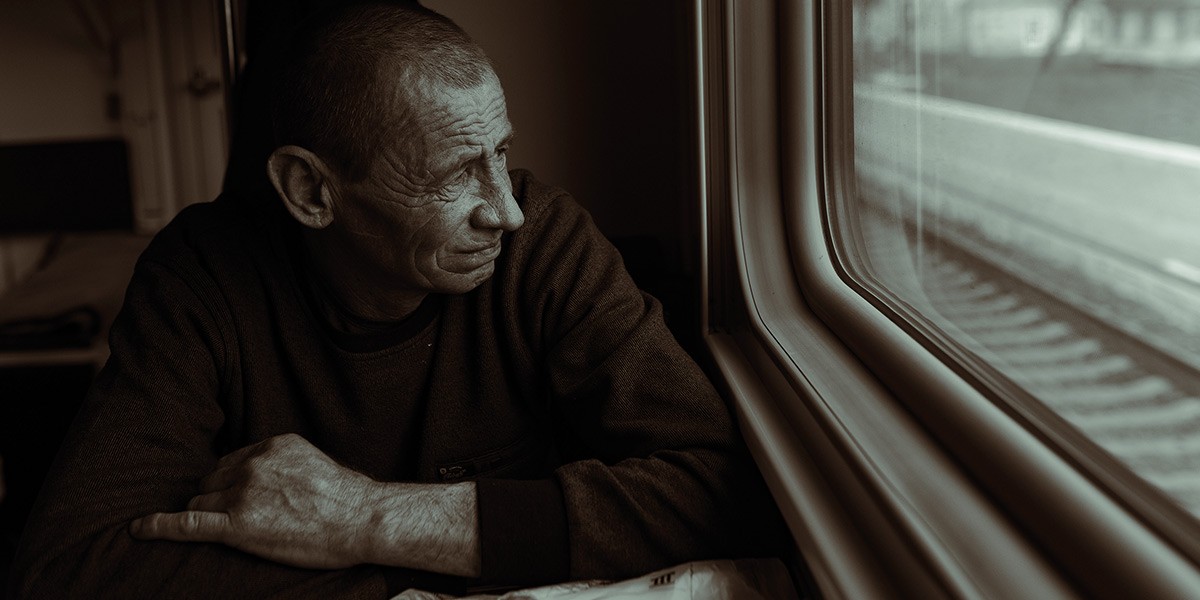 Older man with wrinkles looking pensively out a train window at passing tracks.