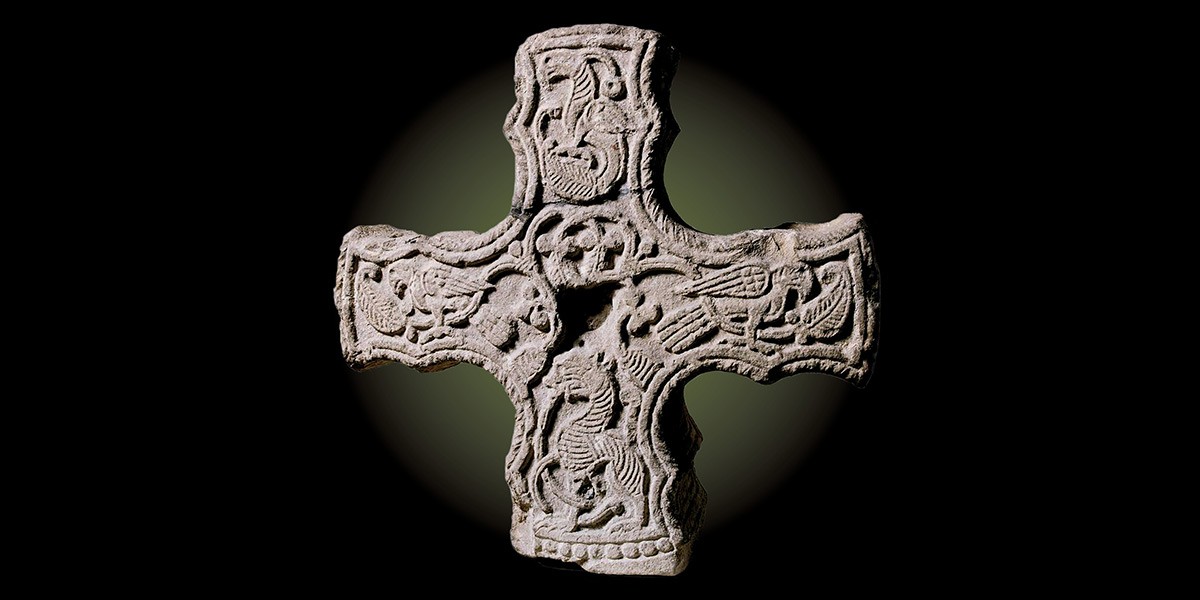 Stone cross with intricate carvings of animals and mythical creatures on a dark background.