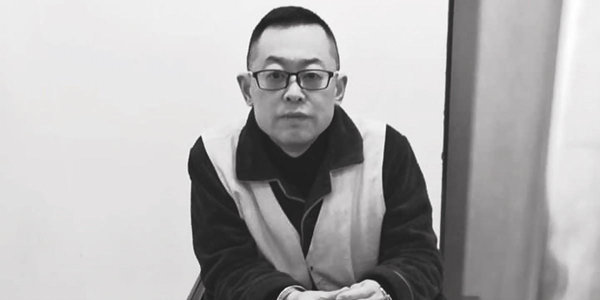 Black-and-white photo of a man with glasses sitting with hands folded, wearing a jacket with light front panels.