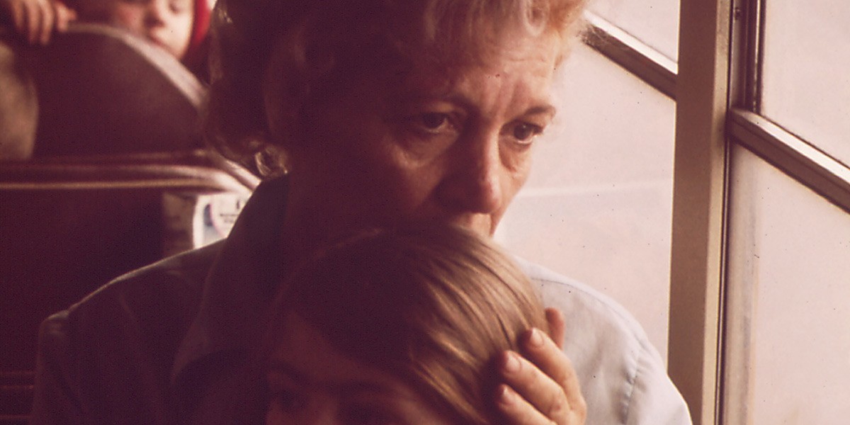 Older woman embraces a young girl on a bus, both looking pensively out the window.