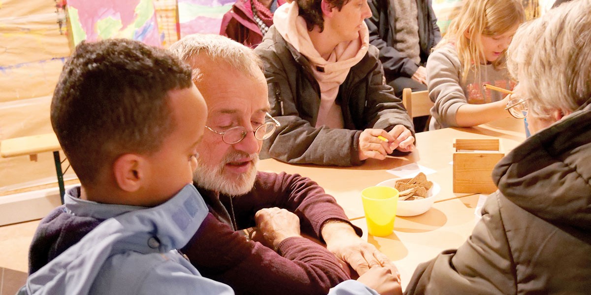 Diverse group including child, elderly man, and adults sitting at table doing crafts with pencils and papers.