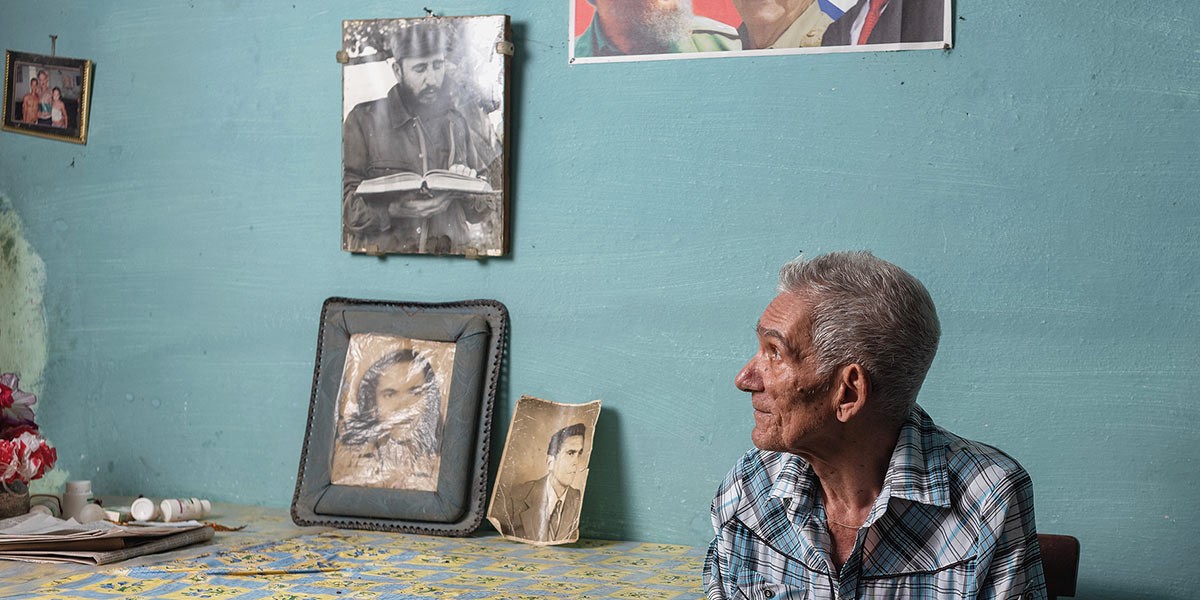 Elderly man in a plaid shirt sitting indoors looking towards a wall with framed old photos and a table.