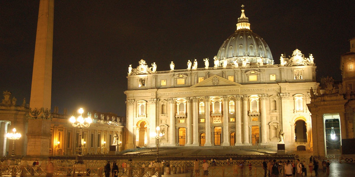 St. Peter's Basilica illuminated at night with statues on top and people in the foreground.