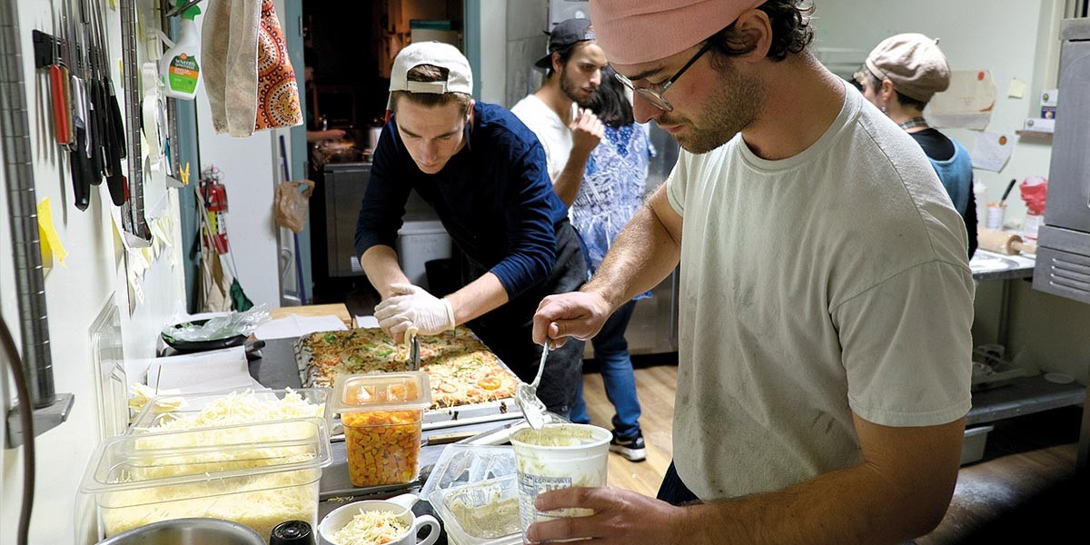Two men prepare pizzas in a kitchen; one adds sauce, and another cuts a large pizza on a tray.