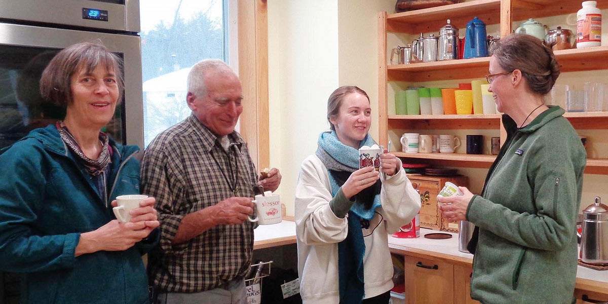 Four people standing in a kitchen, smiling and holding coffee mugs during a casual gathering.