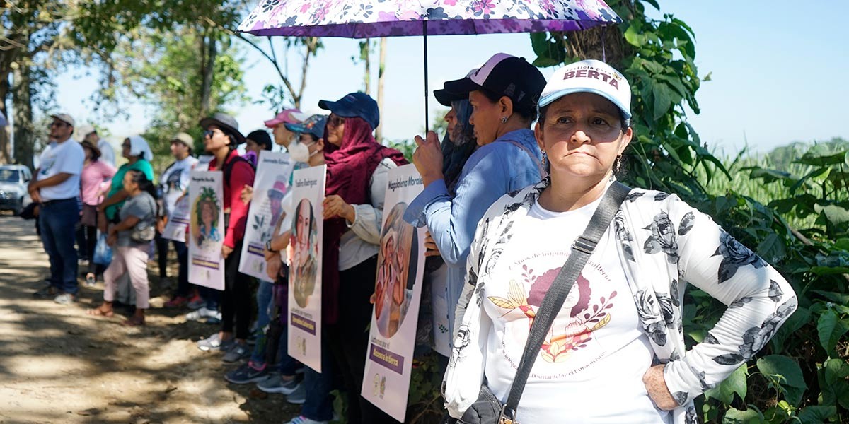 Women standing outdoors in a line holding posters, one woman in front wears a cap that says BERTA.