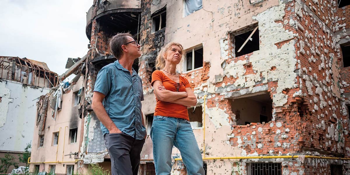Two adults standing with arms crossed, looking at a heavily damaged building with broken walls and windows.