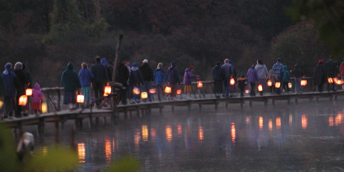 People holding lanterns walking on a curved wooden bridge over misty water at dusk.