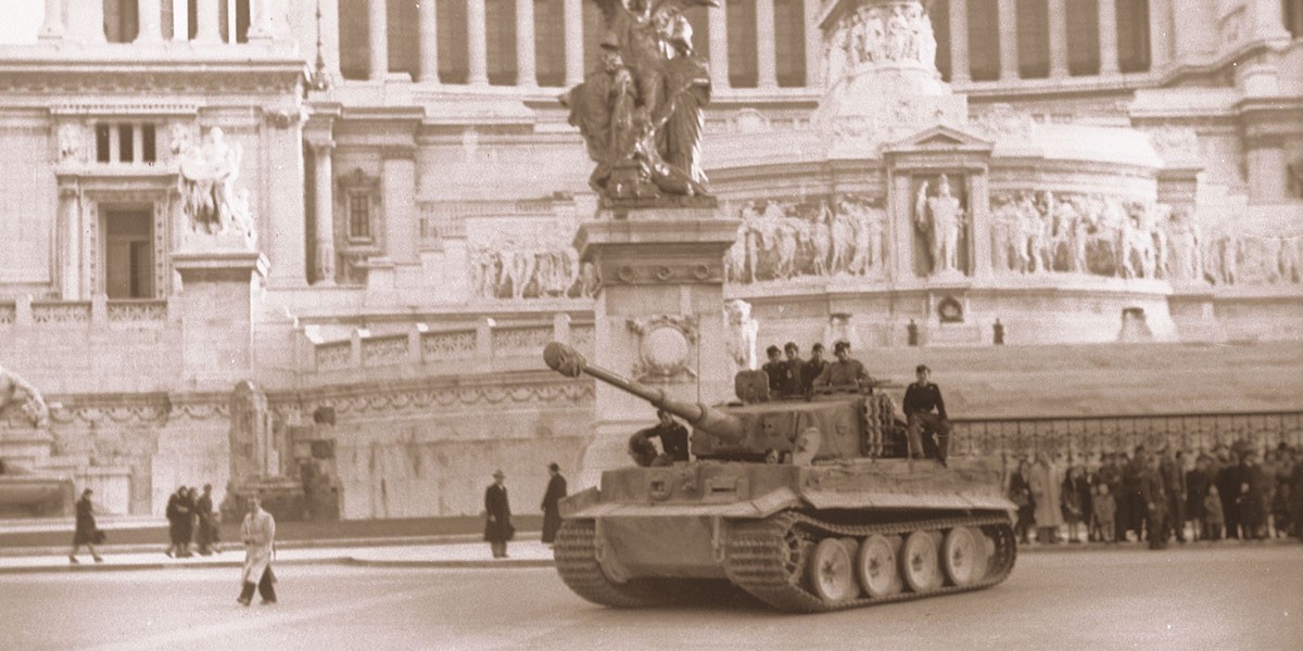 World War II tank with soldiers on it in front of a historic stone monument with sculpted figures.