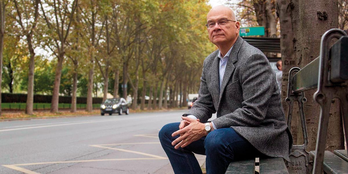 Older man with glasses in a gray blazer sitting on a bench on a tree-lined city street.