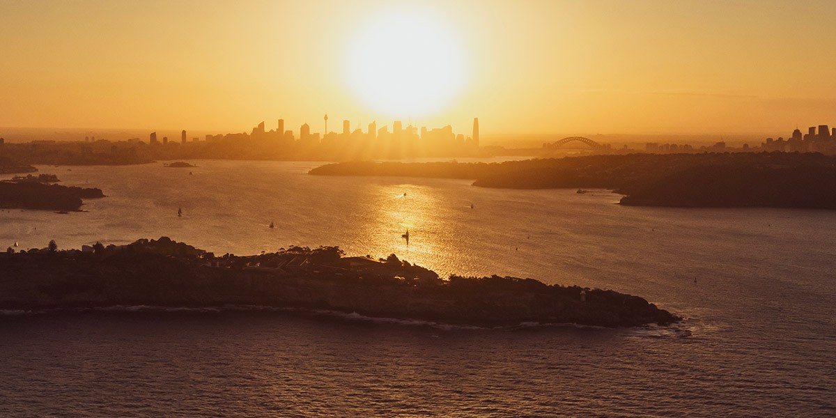 Sunset over a river with city skyline and bridge in the distance, sun reflecting on water surface.