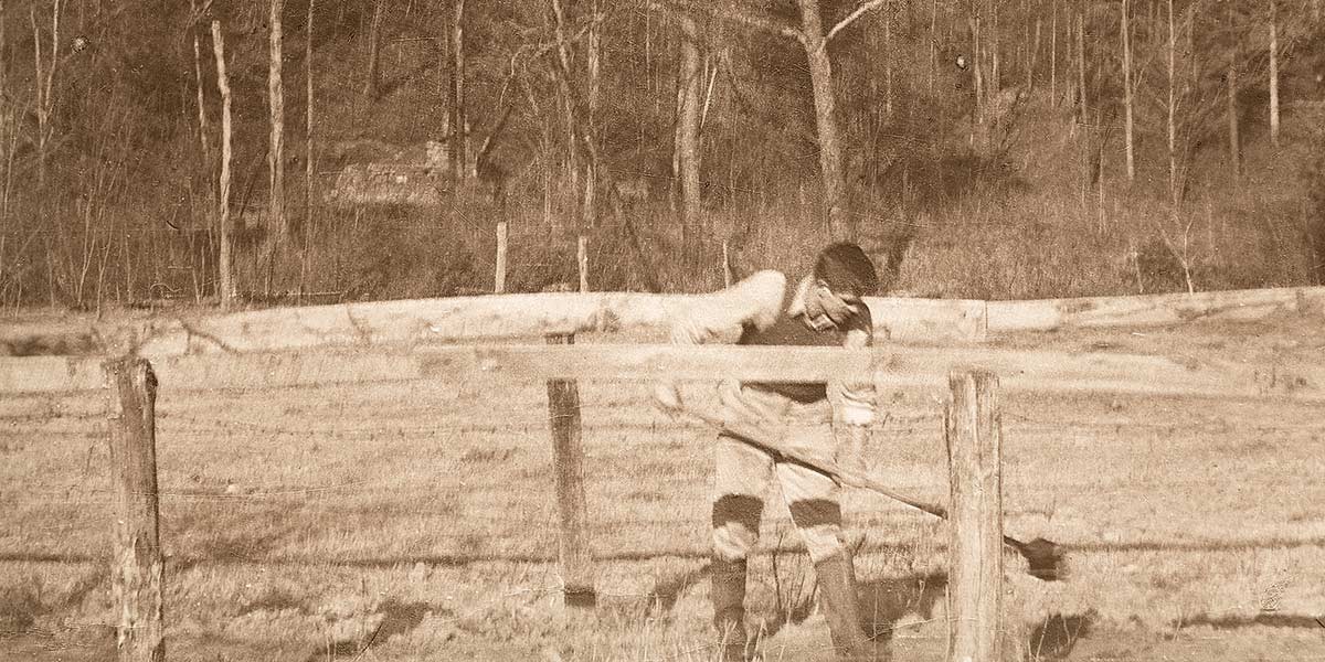 Sepia photo of a man fixing or tending a wooden fence in a rural field with trees in the background.