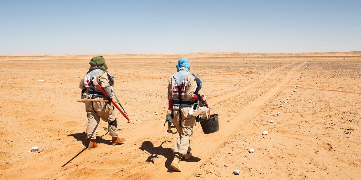 Two people in protective gear walking in a desert clearing marked with small white flags.