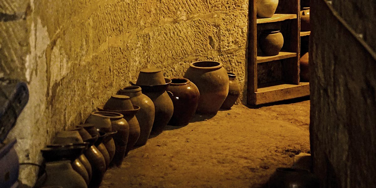 Earthenware pots of various sizes lined up against a textured stone wall on a dirt floor.