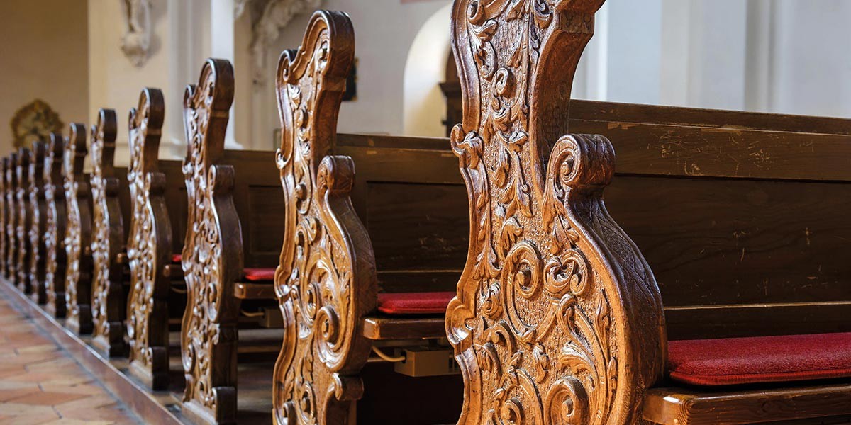 A row of ornately carved wooden church pews with red cushions on a tiled floor.