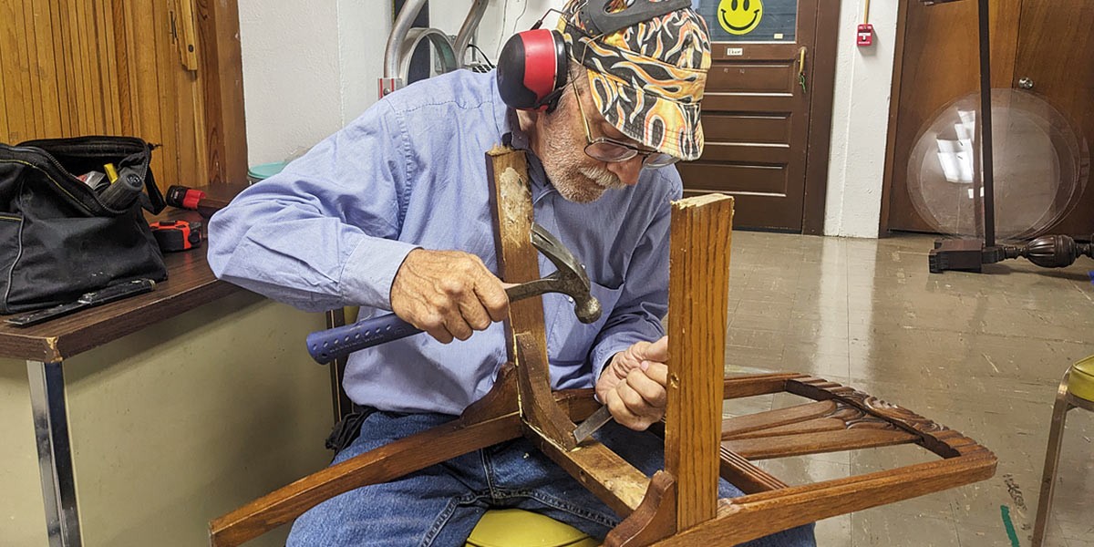 Man wearing hearing protection and cap repairs an upside-down wooden chair using a hammer and chisel.
