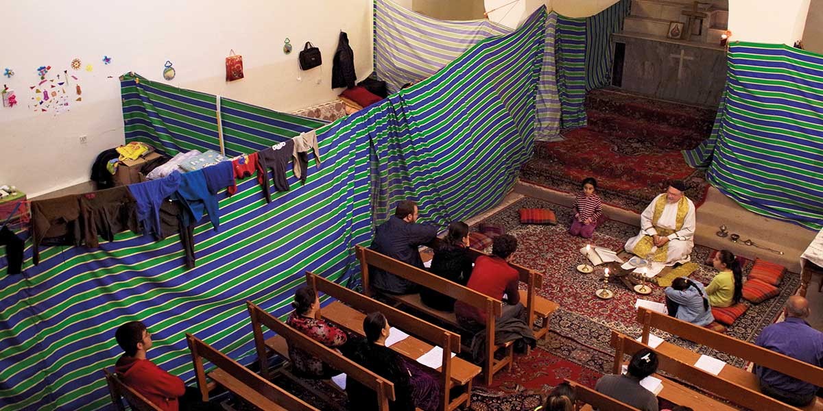 People attending a religious ceremony with a priest seated on a carpeted floor in a small room.