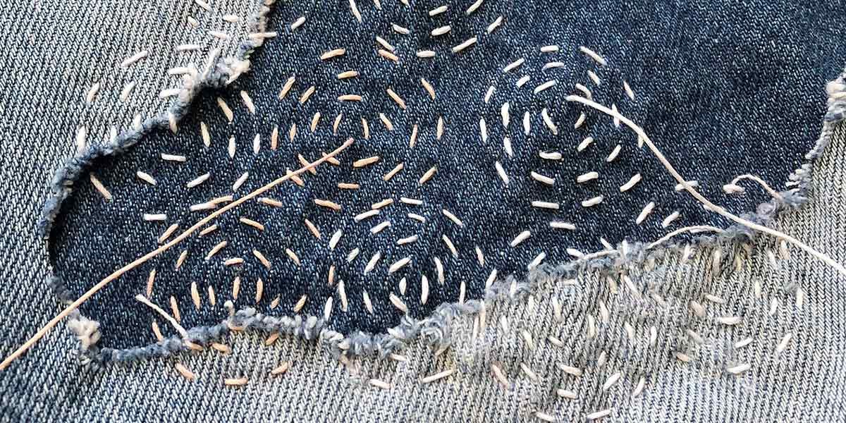 Close-up of blue denim fabric patch sewn with beige running stitches and frayed edges.