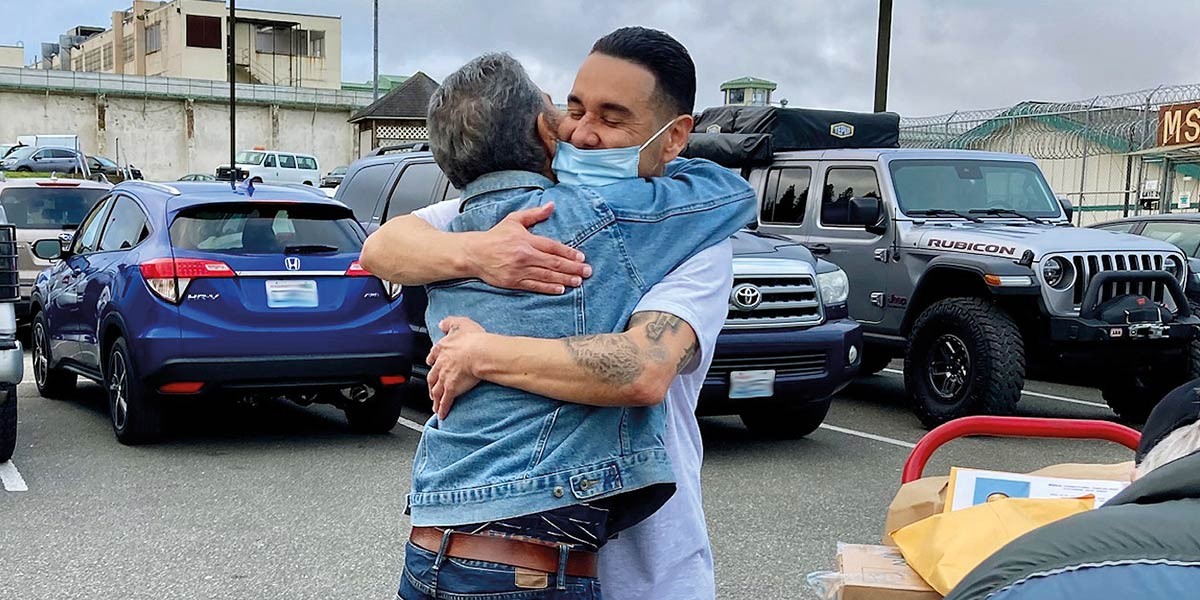 Two men warmly hugging in a parking lot, one wearing a face mask and denim jacket.