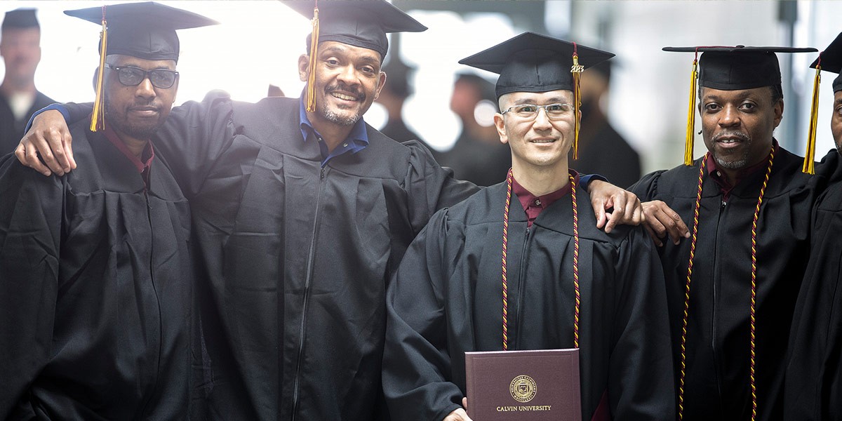 Four male graduates in caps and gowns smiling with one holding a Calvin University diploma.