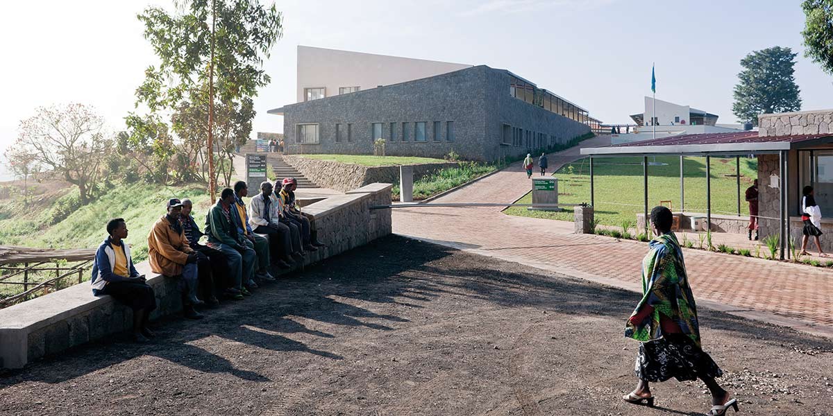 Group of people sitting outdoors on a stone ledge near modern buildings and greenery in daylight.