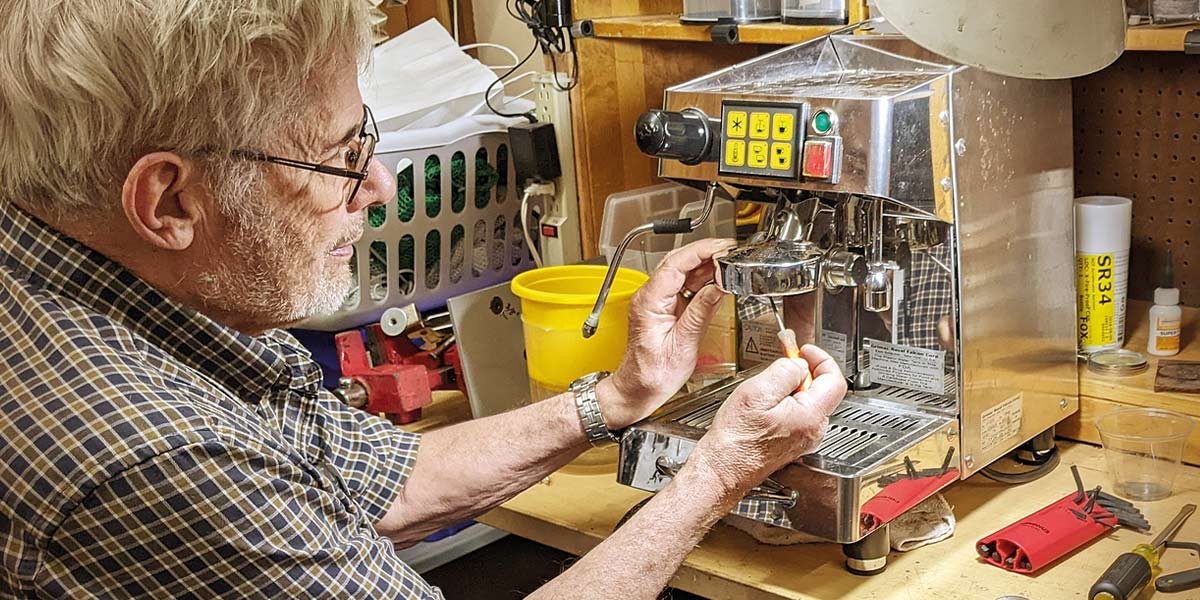Man adjusts parts on a stainless steel espresso machine on a workbench with tools nearby.