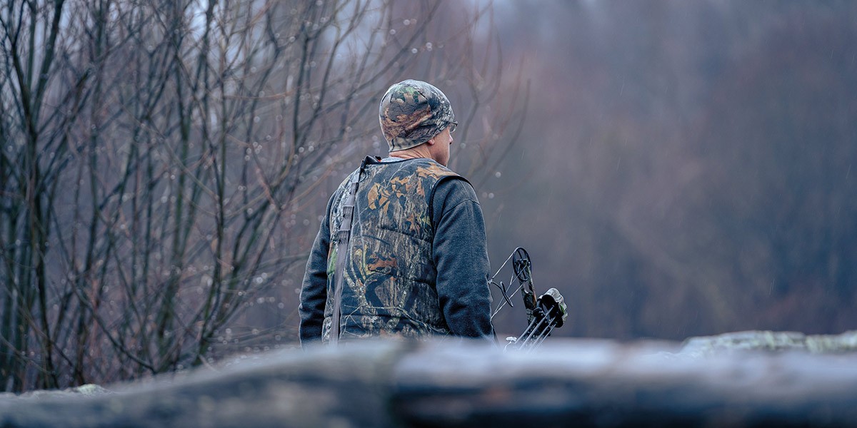 Hunter wearing camouflage gear and hat holding a compound bow in a misty forest.