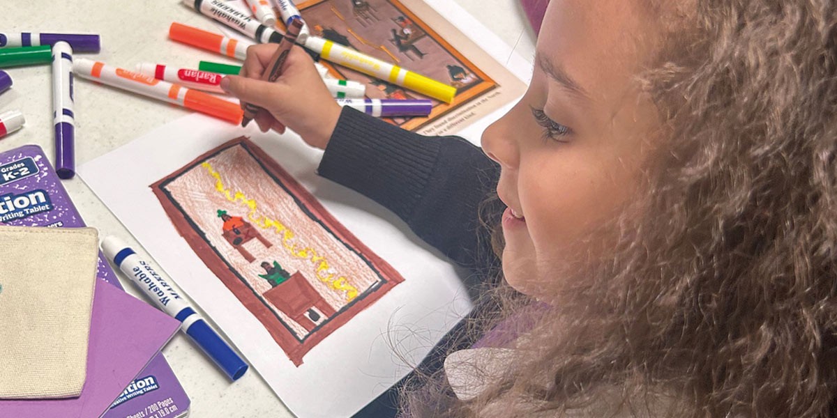 Young girl with curly hair coloring a rectangular drawing with markers on a white table.