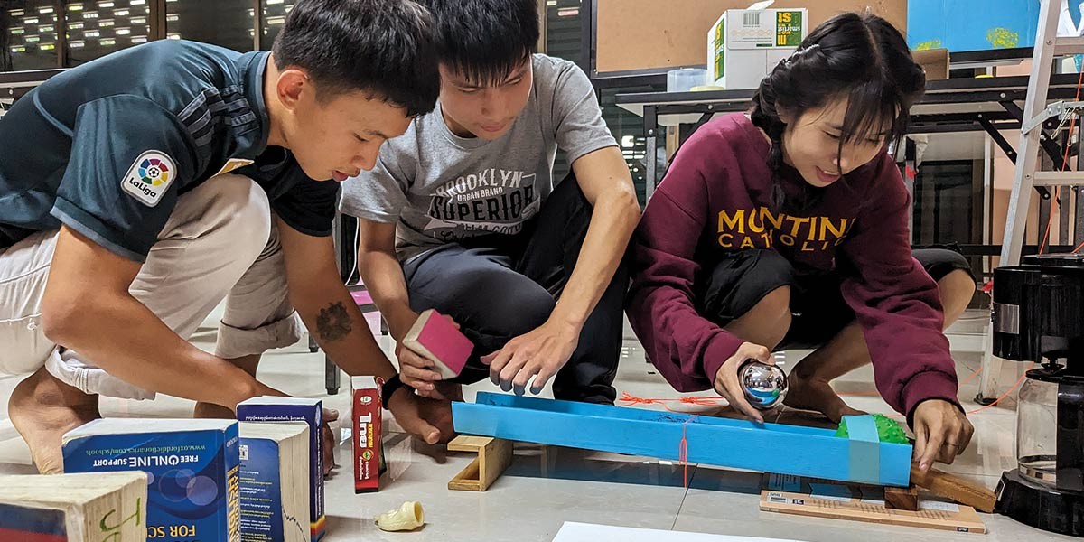 Three young people crouch around a blue ramp holding objects, appearing to conduct a physics experiment.