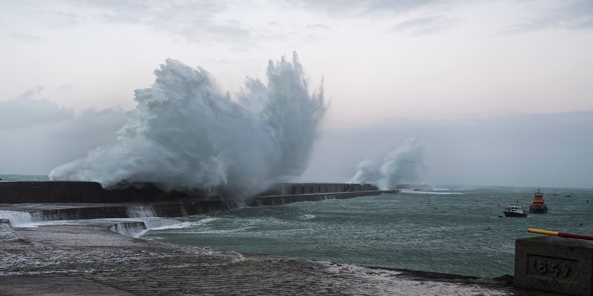 Large waves crashing over a stone pier into a turbulent sea under a cloudy sky.