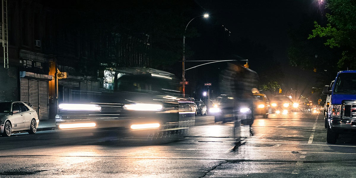 Blurred car lights and a moving pedestrian on a city street at night with parked cars and streetlights.