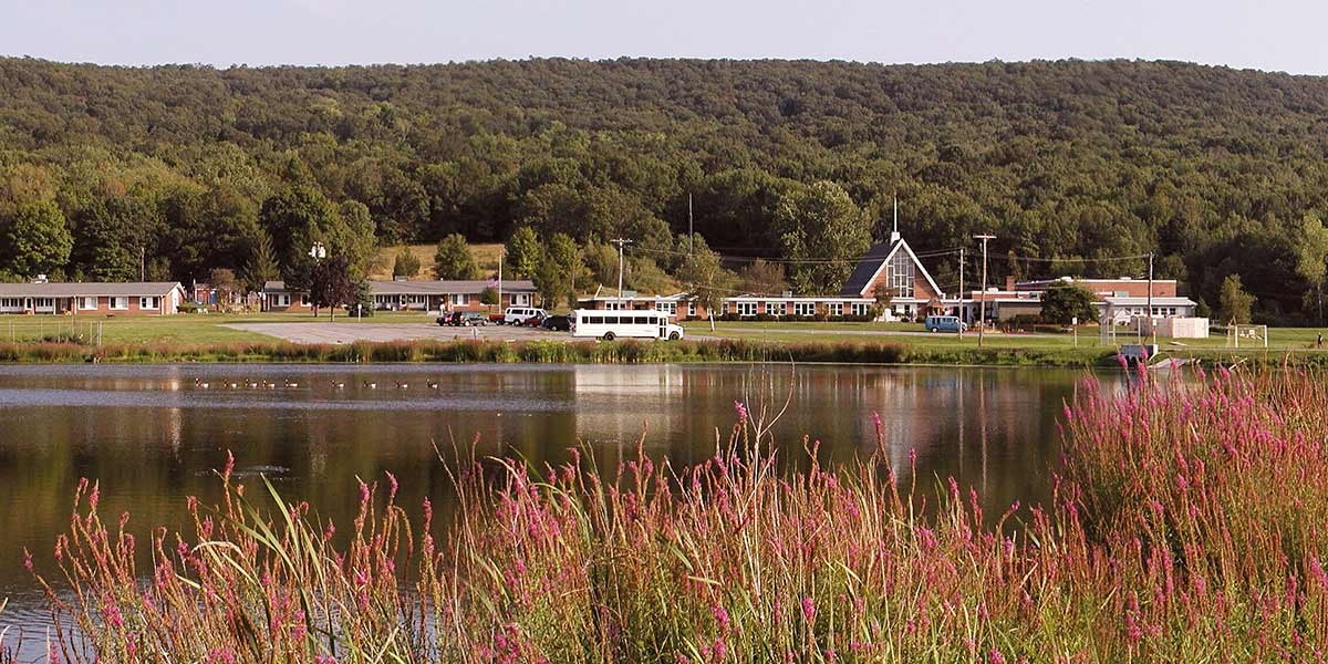 Lake with pink wildflowers in foreground and buildings plus forested hill in background.