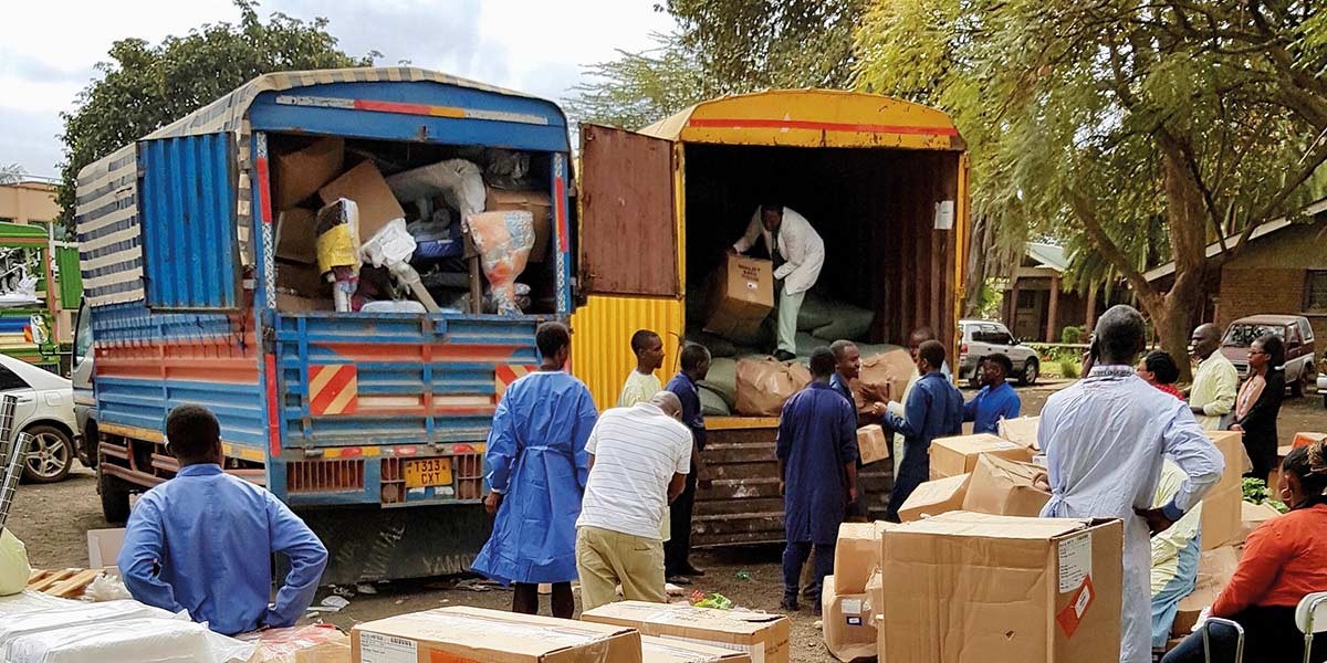Several people unloading large cardboard boxes from two open trucks in an outdoor setting with trees.