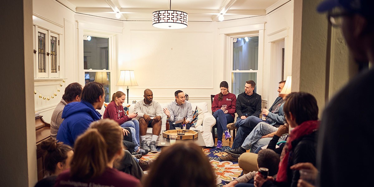 Group of people sitting in a warmly lit living room engaged in conversation and holding drinks.