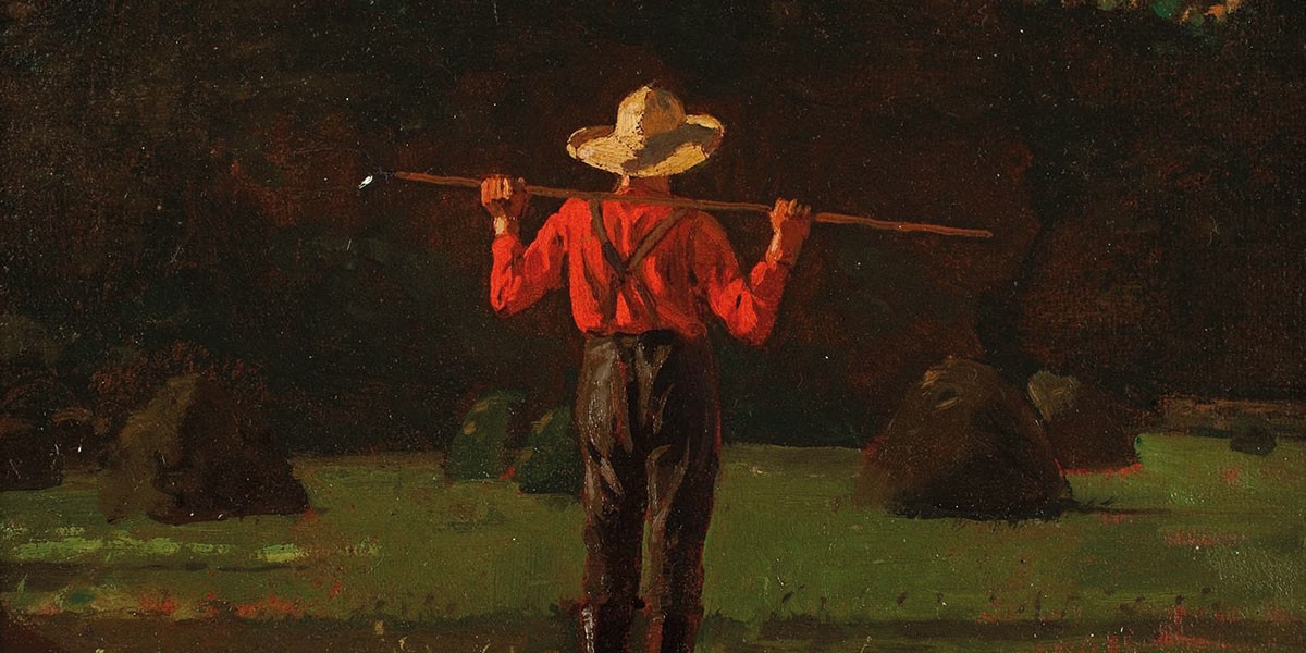 Man in red shirt and straw hat standing in a field holding a long stick across his shoulders.