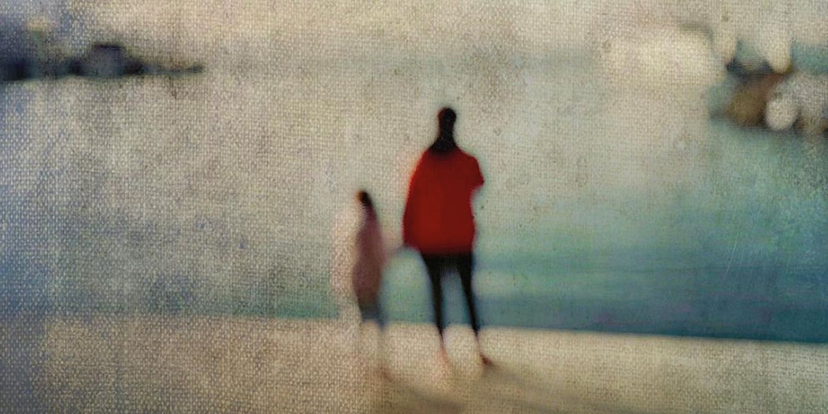 Blurry image of two people standing near a body of water, one wearing a red top.