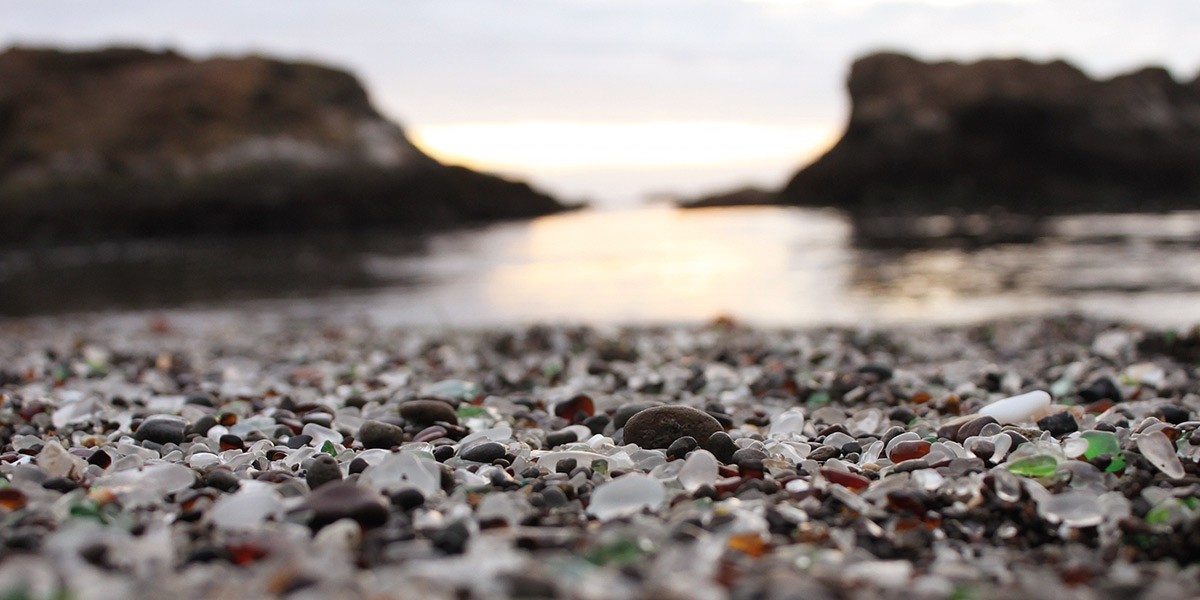 Close-up of sea glass and pebbles on a beach with blurred rocky coastline in background at sunset.