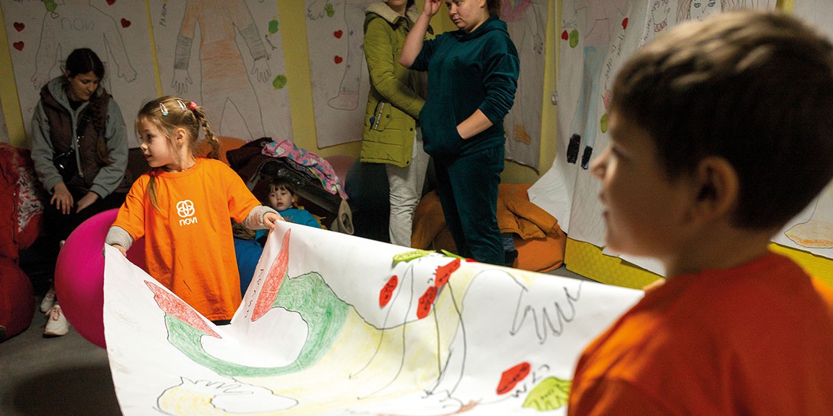 Two children in orange shirts holding a large colorful drawing in a room with people and wall art.