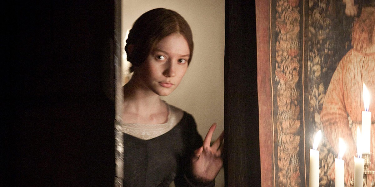 Young woman in period dress peeking around a door, with lit candles and tapestry in the background.