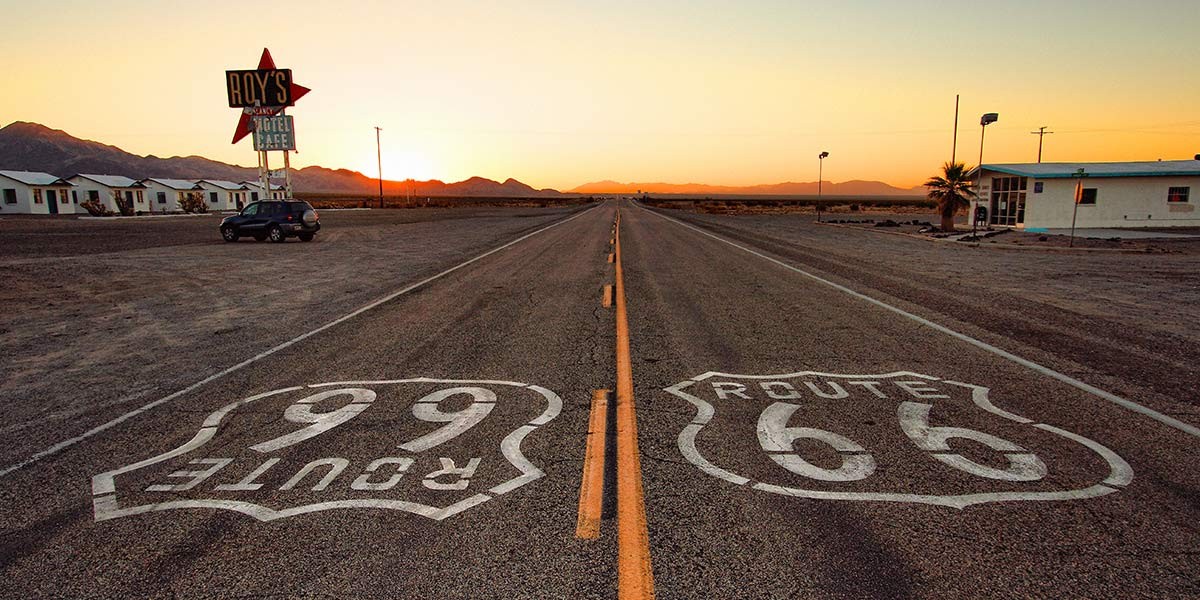 Empty Route 66 and Route 99 roads stretch straight towards a sunset with Roy's Motel and Cafe sign on left.